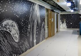 A hallway with dark walls featuring white abstract art resembling stars and celestial objects. Wooden doors line one side.