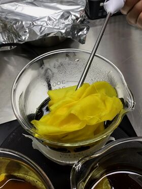Keltainen kangas sekoitetaan lasimaljassa, joka on täynnä tummaa nestettä. Taustalla on alumiinifoliota.
