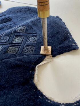 Metallityökalu, jossa on puinen kahva, painaa tummansinistä kangasta, jossa on ommeltuja kuvioita, leikaten sitä.