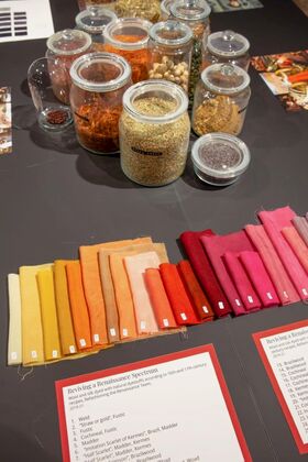 Lasipurkkeja, joissa on erilaisia luonnonväriaineita, ja kangasnäytteitä keltaisissa, oransseissa, punaisissa ja vaaleanpunaisissa sävyissä pöydällä.