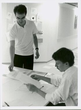 Kaksi ihmistä työskentelee pöydän ääressä, yksi seisoo ja yksi istuu, huoneessa on papereita ja piirustuksia seinällä.