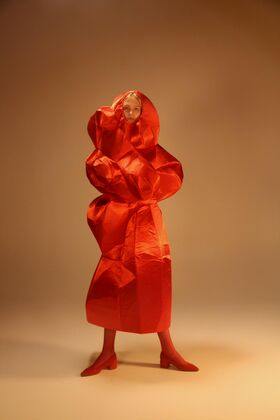 Henkilö, jolla on ylisuuria, ryppyinen punainen asu ja punaiset kengät, seisoo beige taustaa vasten.