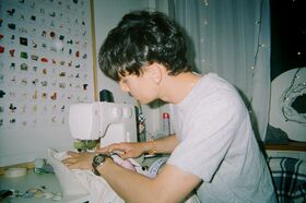 A person sewing with a white sewing machine in a room with a poster of chairs and fairy lights.