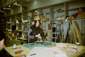 A person wearing a helmet works with tools at a cluttered workshop table. Shelves filled with various items are in the background.
