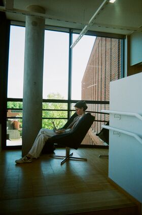 Person sitting in a chair by a large window, reading from a laptop. Brick building and trees visible outside.