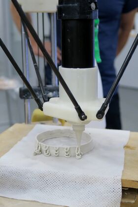 A 3D printer extruding a circular object on a white mesh surface. A person is visible in the background.