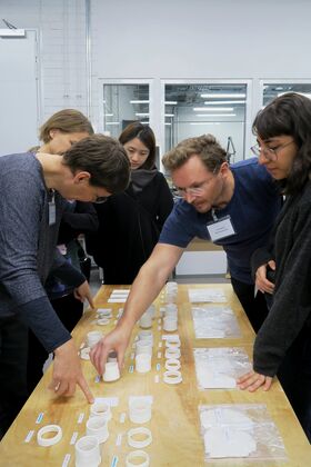 People examining various white cylindrical objects and samples on a wooden table in a bright room.
