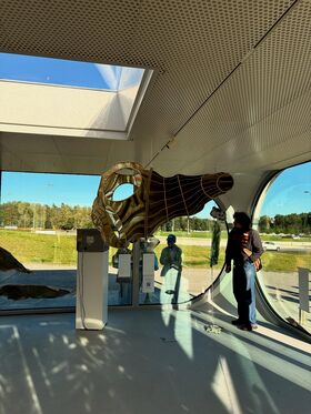 A person stands inside a modern building with large windows, observing a hanging sculpture. Outside, people walk by.