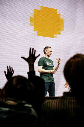 Henkilö vihreässä paidassa seisoo lavalla, yleisössä kädet ylhäällä. Taustalla ruudulla pikselöity keltainen kuvio.