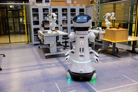 En humanoid robot i ett laboratorium med robotarmar och arbetsstationer i bakgrunden.