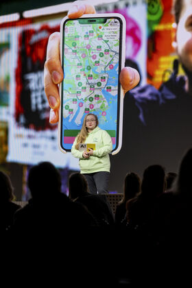 Henkilö vihreässä hupparissa esittelee suuren näytön edessä, jossa näkyy Helsingin kartta älypuhelimessa.