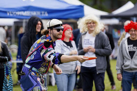 A person in a colourful outfit throws a frisbee at an outdoor event. Others stand in the background under tents.