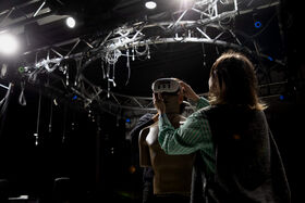 A person in a green striped shirt adjusts a device on a mannequin in a room with many cables and lights.