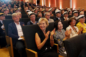 A large audience in an auditorium, many wearing white caps, clapping. Front row has people in formal attire.