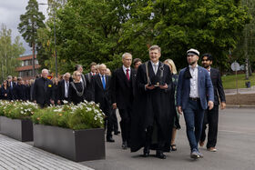 A procession of professors in formal attire walks outside. In the foreground, the rector and student union president with a doctoral hat.