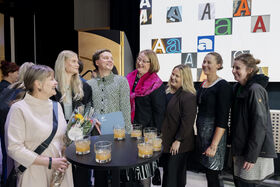A group of people standing around a table with drinks. A wall with various letter 'A' designs is in the background.