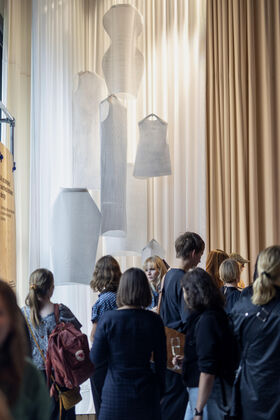 Ihmiset tarkkailevat valkoisia tekstiiliveistoksia, jotka roikkuvat beigejen verhojen edessä.