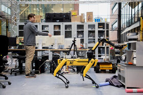 En person interagerar med en gul Boston Dynamics robot hund i ett rörigt kontorsutrymme med olika utrustning och hyllor.