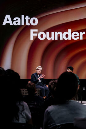 Henkilö tummassa puvussa puhuu lavalla, taustalla suuri näyttö, jossa lukee 'Aalto Founder'.