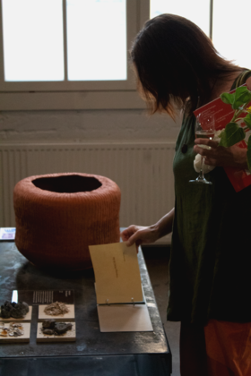 A person in a green top and orange trousers examines a booklet near a large textured clay urn, holding a glass and a red catalogue.