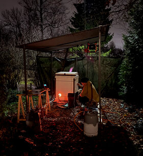 A person in a yellow jacket works with a kiln under a canopy at night. Trees and bushes surround the area.