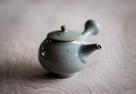 A small, round, green ceramic teapot with a short spout and a lid, placed on a light wooden surface.