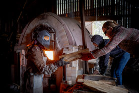 Two people are placing wood into a large brick kiln inside a wooden structure. One person wears protective gear.