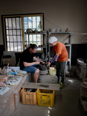 Two people working on pottery in a workshop. Shelves with ceramic items and a window are in the background.