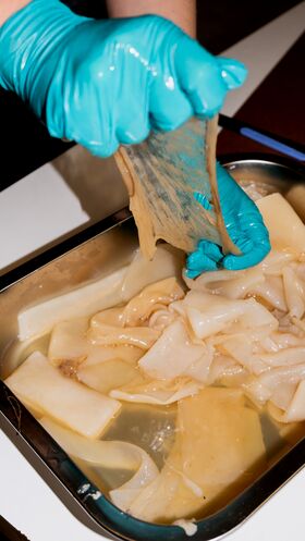 A person wearing blue gloves handles a translucent sheet in a tray filled with similar sheets submerged in liquid.