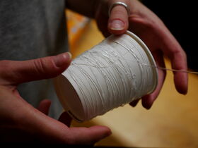 Hands holding a white paper cup wrapped with clear string. The background is blurred and features warm tones.