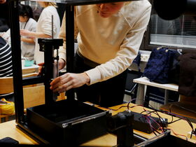 A person adjusts a syringe on a 3D printer in a workshop setting. Other people are working in the background.