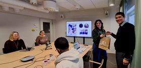 A group of people in a meeting room with a presentation screen showing images. One person is handing a paper bag to another.