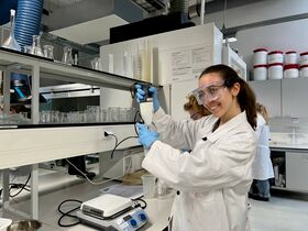 A person in a lab coat and blue gloves uses a handheld device in a laboratory with various glassware and equipment.