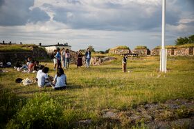 People are gathered outdoors on a grassy field with stone structures in the background under a cloudy sky.