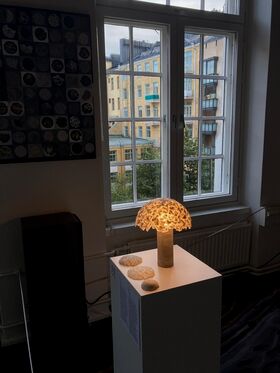 A glowing lamp with a textured shade on a pedestal near a window. Buildings and trees are visible outside.