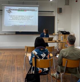 A classroom with a presentation screen showing a research title. Three people are seated, facing the presenter at a desk.
