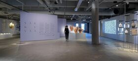 A modern art gallery with pottery, hanging art, and a projection. A person walks past a lavender wall with text.