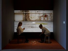Two people sit facing each other in a room with a projected image of an empty chair in a dilapidated space.