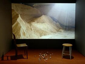 A room with a chair, a stool, and scattered stones on the floor. A large photo of sand dunes is displayed on the wall.