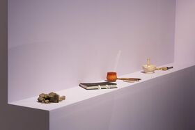 A white shelf with rocks, a book, a clay pot, brushes, and a mortar and pestle against a light purple wall.