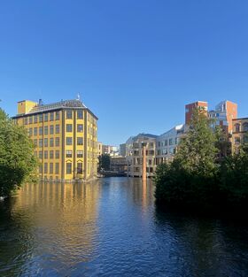 A yellow building by a river, surrounded by modern buildings and trees under a clear blue sky.