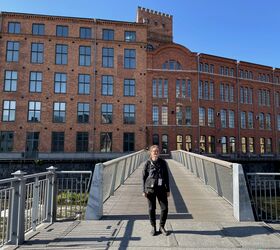 A person stands on a bridge in front of a large red-brick building with many windows on a sunny day.