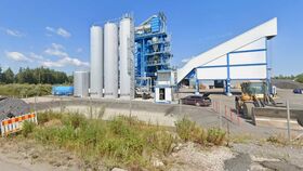An industrial site with tall silver tanks, blue structures, a bulldozer, and a fenced area. Trees are visible in the background.
