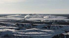 Snow-covered industrial landscape with smoke rising from chimneys, surrounded by hills and distant forest.
