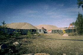 Construction site with sand piles, machinery, and graffiti-covered walls. Trees and rocks in the foreground.