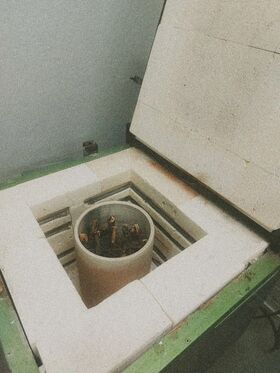 An open kiln with a cylindrical container inside, holding small pieces of wood. The kiln has a green exterior.