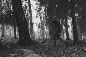 A person in camouflage gear runs through a dense forest with tall trees and fallen leaves on the ground.