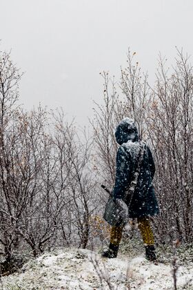 Person in a blue coat and yellow patterned trousers standing in a snowy forest, holding a shovel.