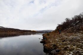 Two people near a calm river, one crouching by the water, the other walking on a rocky, snow-dusted shore under a cloudy sky.