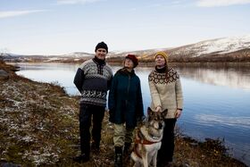 Three people and a dog stand by a lake with snowy hills in the background. They wear winter clothing.